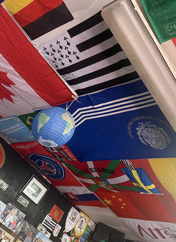 A room with a ceiling covered in international flags, including those of Canada, Germany, China, Sweden, and the black-and-white Breton flag. A small hanging globe lantern is suspended from the ceiling. The walls below are decorated with photos, patches, and additional flags