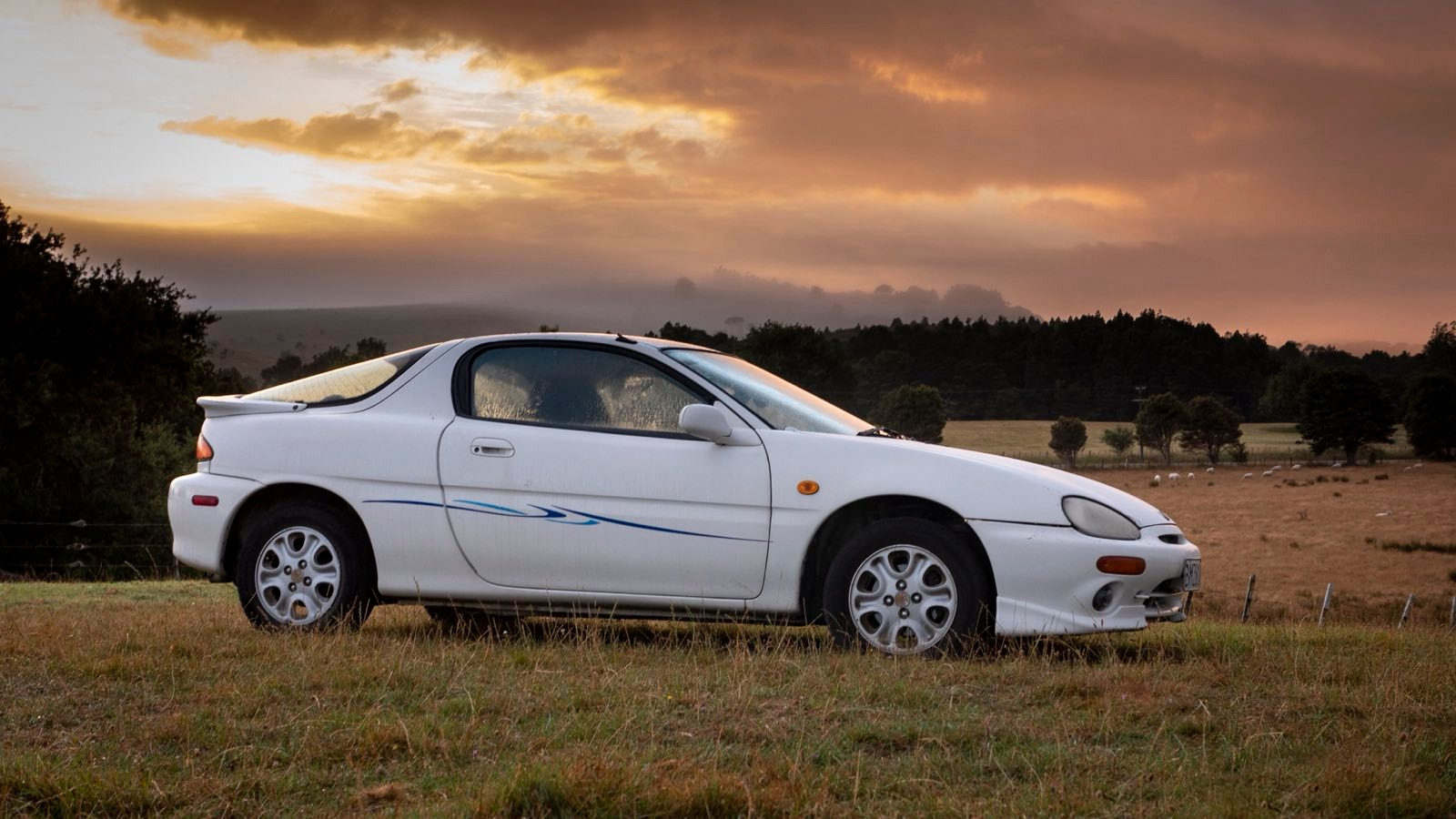 My first car. A white, two door sports car. It has a weird blue 80s/90s wave pattern on the side. The picture is taken on a grassy hill. In the background there is farmland, with some sheep visible. The sky is gold, it was taken in the morning, or in the evening.